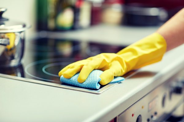 people, housework and housekeeping concept - close up of woman hand in protective glove with rag cleaning cooker at home kitchen