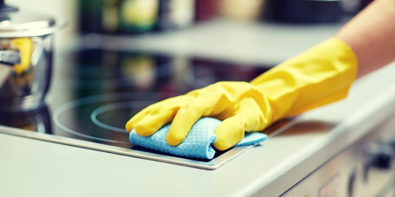 people, housework and housekeeping concept - close up of woman hand in protective glove with rag cleaning cooker at home kitchen